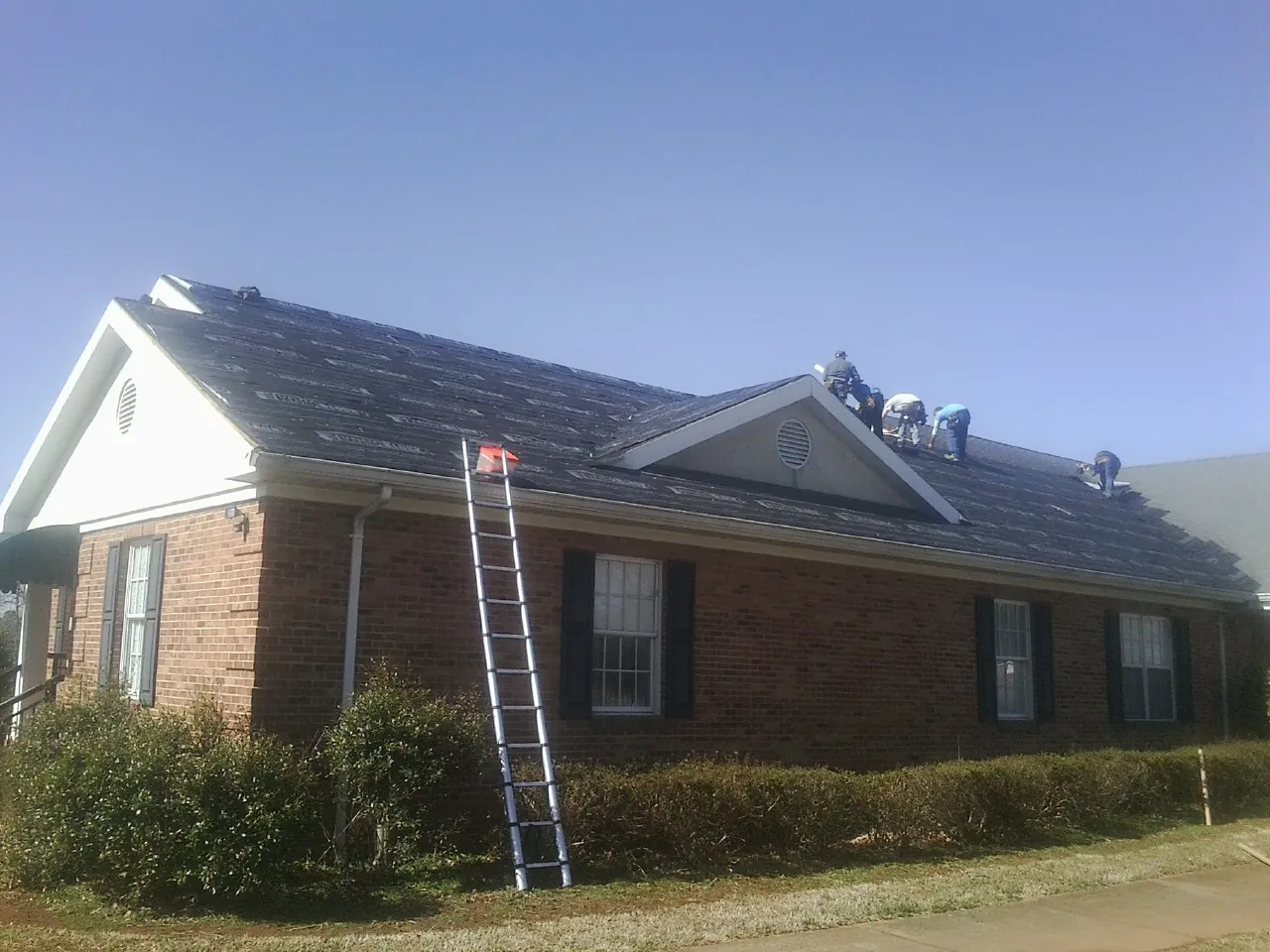 Crew tearing shingles off a brick ranch roof with a ladder set — Emergency Roof Repair work by our crew in Belleview