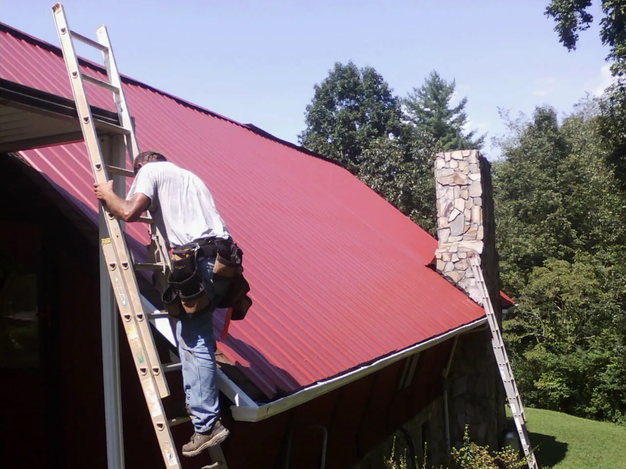 Roofer on a ladder beside a red metal roof and stone chimney — Soffit & Fascia Repair by Roofing Brothers in Belleview
